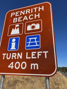 Brown tourist direction sign indicating Penrith Beach with icons for attractions, information, and picnic facilities, advising motorists to turn left in 400 metres.